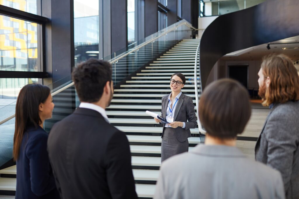 A young woman wearings a dress suit and skirt stands in front of a long stairway talking as others watch. Seems to be providing a tour.