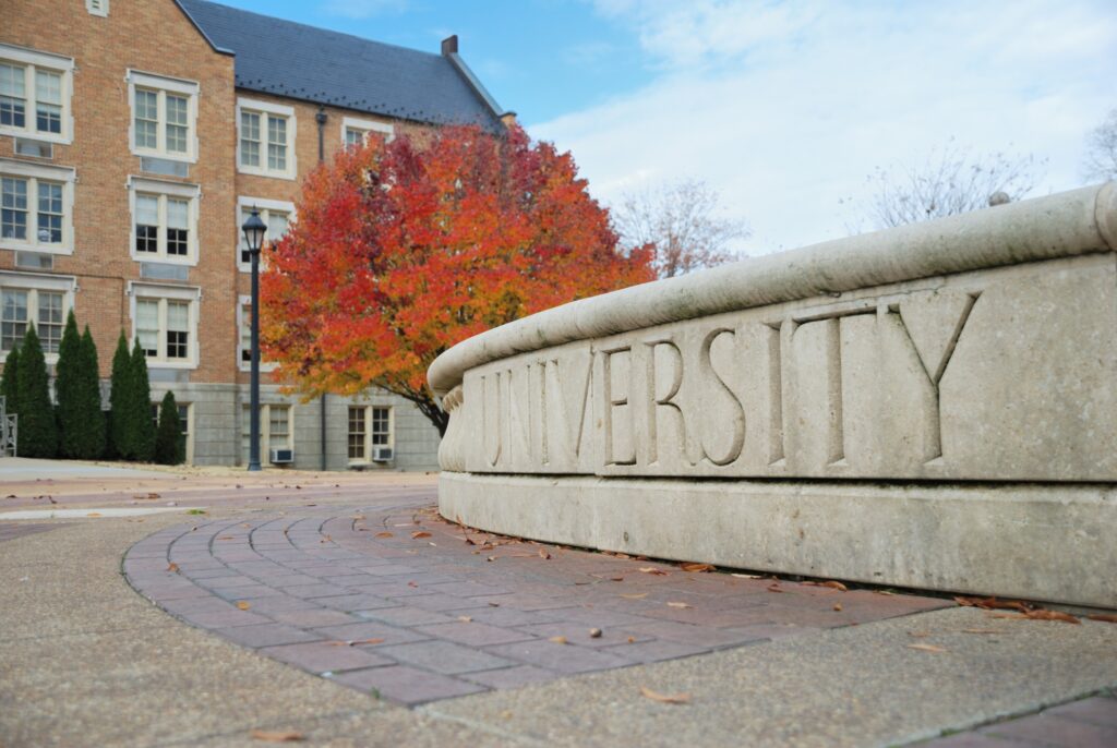 A curved wall with the word university engraved into it. Beyond it is a tall older building and a bright orange-red tree.